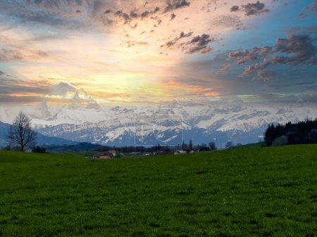 Impresionante puesta de sol sobre un exuberante paisaje suizo, con un sinuoso camino a través de campos verdes vibrantes que conducen a los majestuosos Alpes nevados, bajo un cielo dramático pintado con tonos de naranja y azul