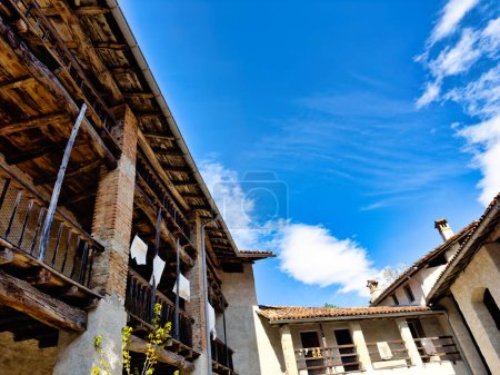 Casa de campo tradicional europea con un patio abierto, con múltiples niveles de balcones, vigas a la vista y viejos carros agrícolas de madera, frente a un cielo azul brillante