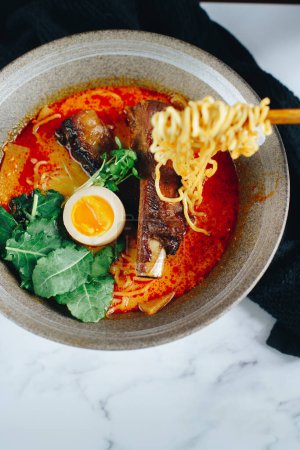 A bowl of ramen with a piece of egg in it. The noodles are long and the broth is red