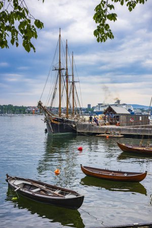 Foto de Oslo, Noruega - 21 de junio de 2023: Las embarcaciones atracadas utilizadas en la pesca, descansan a lo largo de las costas de la Península de Bygody y se encuentran en el Oslofjord Interior, ya que la ciudad de Oslo sirve como fondo durante un día nublado.. - Imagen libre de derechos
