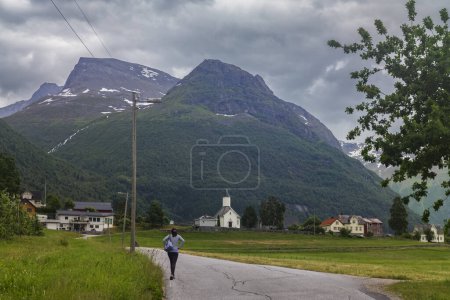 Téléchargez les photos : Loen (Norvège), le 26 juin 2023 : Une femme se rend à l'église de Loen, une église paroissiale de l'église de Norvège, photographiée ici de face, par une soirée nuageuse. - en image libre de droit