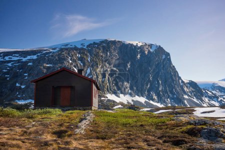 Téléchargez les photos : La neige a recouvert la vallée de Breiddalen au parc national de Jotunheimen en Norvège, le long de la route de Geiranger, pendant une journée d'été. Une cabane repose près du pied d'une montagne. - en image libre de droit