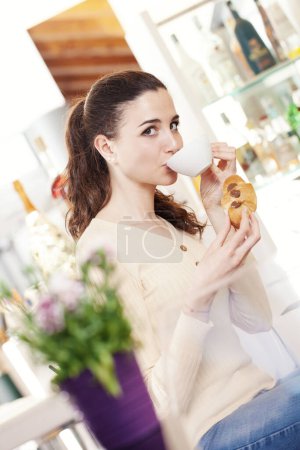 Foto de Hermosa joven sonriente con croissant y una taza de café en sus manos en la cafetería, heladería y pastelería. Retrato de chica morena bebiendo y mirando a la cámara. - Imagen libre de derechos