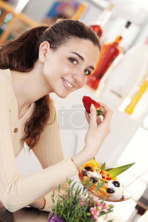Foto de Hermosa joven sonriente con taza de ensalada de frutas con helado en la parte superior en la cafetería, heladería y pastelería. Retrato de niña comiendo helado, fruta y fresa. - Imagen libre de derechos