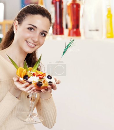 Foto de Hermosa joven sonriente con taza de ensalada de frutas con helado en la parte superior en la cafetería, heladería y pastelería. Retrato de chica morena riendo y mirando a la cámara - Imagen libre de derechos