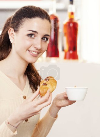 Foto de Hermosa joven sonriente con croissant y una taza de café en sus manos en la cafetería, heladería y pastelería. Retrato de chica morena riendo y mirando a la cámara. - Imagen libre de derechos