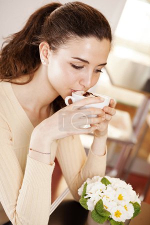 Foto de Hermosa joven bebiendo una taza de café en sus manos en la cafetería, heladería y pastelería. Retrato de morena bebiendo un capuchino cerca de las flores. - Imagen libre de derechos