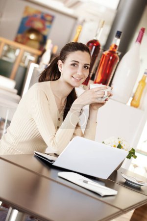 Foto de Hermosa joven sonriente bebiendo una taza de café en la cafetería, heladería y pastelería. Estudiante chica con portátil mirando a la cámara. - Imagen libre de derechos