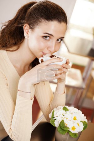 Foto de Hermosa joven sonriente bebiendo una taza de café en la cafetería, heladería y pastelería. Ella disfrutando de su capuchino. Retrato de chica morena mirando a la cámara - Imagen libre de derechos