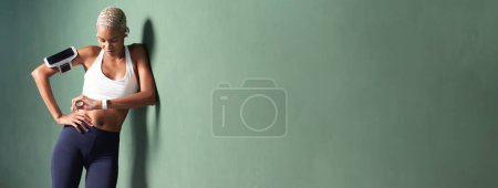 Foto de Deportista mujer atleta corriendo entrenamiento y correr, sonriente mujer afroamericana mirada reloj inteligente fitness, lleva auriculares y teléfono inteligente en el brazalete. Apoyado en una pared verde urbana con espacio para copias - Imagen libre de derechos
