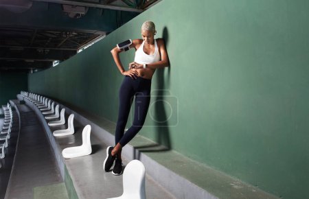 Foto de Deportista mujer atleta durante el entrenamiento de carrera y correr, sonriente mujer afroamericana mirar un reloj inteligente fitness, usar auriculares y smartphone a brazalete. Utilización de gradas de estadio vacías y escaleras - Imagen libre de derechos
