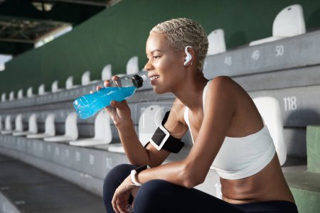 Foto de Una atleta sonriente bebiendo bebida energética de la botella después de hacer ejercicio deportivo, usa auriculares y reloj inteligente. Mujer afroamericana sentada en estadios vacíos y escaleras - Imagen libre de derechos
