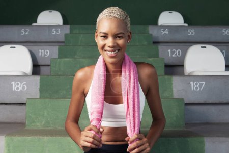 Foto de Atleta sonriente después de correr ejercicio deportivo de entrenamiento, con toalla. Mujer afroamericana sentada en estadios vacíos y escaleras - Imagen libre de derechos