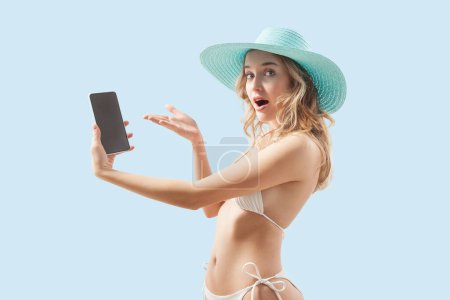 amazed woman dressed in a white bikini an big blue sun hat is showing a smartphone. Image is isolated on a light blue background concept of online shopping for summer beach holiday or vacation travel
