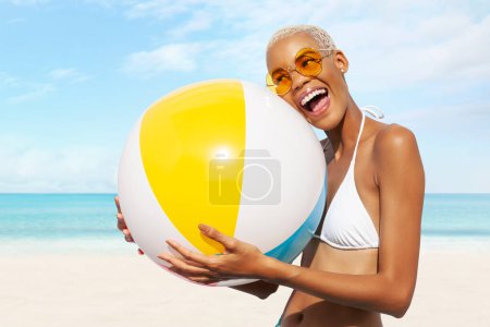 Téléchargez les photos : Fille heureuse sur le côté de la plage tient ballon de plage gonflable, portant des lunettes de soleil et bikini se sent bien, femme afro-latino-américaine. Concept de vacances au bord de la mer ou shopping pour des vacances d'été à la plage - en image libre de droit