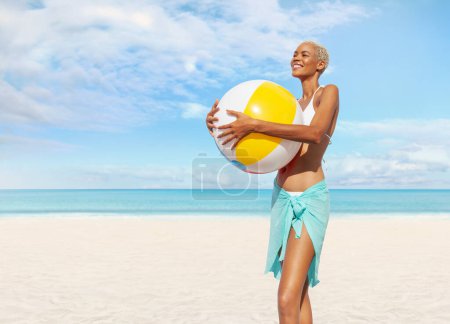 Téléchargez les photos : Fille souriante sur le côté de la plage tient ballon de plage gonflable, portant un bikini et un paréo, femme afro-latino-américaine. Concept de vacances au bord de la mer ou shopping pour des vacances d'été à la plage - en image libre de droit