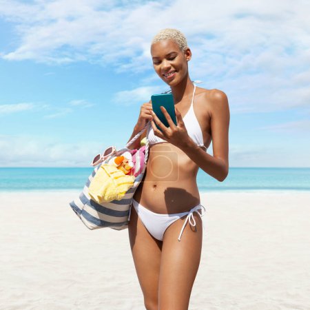 Foto de Mujer feliz en el lado de la playa con bikini sosteniendo una bolsa de playa y el uso de teléfono móvil en un día soleado con cielo azul. Concepto de vacaciones en la playa de verano, compras en línea, reserva de viajes y resort - Imagen libre de derechos