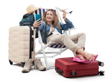 Foto de Mujer turista en traje de viaje, en la silla de cubierta con maletas de carro, mostrar un modelo de avión, utilizando el teléfono móvil. Vacaciones de verano en la playa, reserva de vuelos y vacaciones. Viajes influencer estilo de vida - Imagen libre de derechos