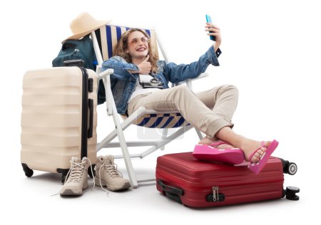 Foto de Mujer turista en traje de viaje, en la silla de cubierta con maletas de carro, hacer selfie con teléfono móvil y pulgares hacia arriba. Vacaciones de verano en la playa, reserva de vuelos y vacaciones. Viajes influencer estilo de vida - Imagen libre de derechos