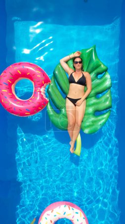 Foto de Caucasian female in a black bikini relaxes in her garden pool on a sunny summer weekend. Beautiful young woman lies on a big leaf floatie in her crystal clear home pool. - Imagen libre de derechos