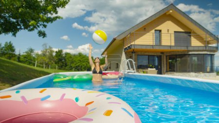 Téléchargez les photos : Playful young woman throws an inflatable ball at other floaties in her garden pool. Caucasian female in black bikini plays with the floaties in her private pool on a sunny summer day - en image libre de droit