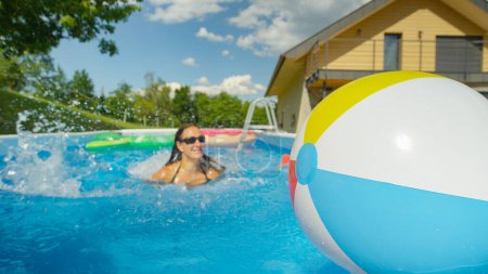 Photo for Smiling woman swims toward an inflatable ball floating around her pool. Young Caucasian female tourist is swimming towards a floating in her home pool in the idyllic green backyard. - Royalty Free Image