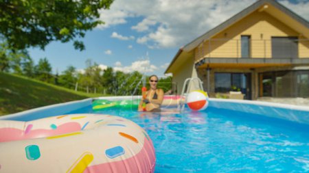 Foto de Happy young woman squirts water with a big plastic water gun while playing in the garden pool behind her modern home. Joyful Caucasian female is enjoying the summertime in her home pool - Imagen libre de derechos