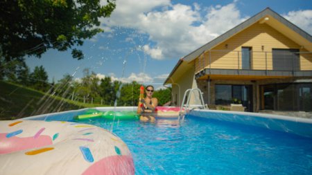 Foto de Joyful Caucasian female is enjoying the summertime in her home pool. Happy young woman squirts water with a big plastic water gun while playing in the garden pool behind her modern home - Imagen libre de derechos
