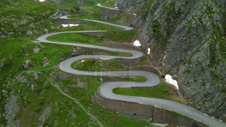 Foto de AERIAL: Vista aérea cinematográfica de un camino de horquilla serpenteando una montaña en Suiza. Turistas en coche blanco se detienen para observar los pintorescos Alpes suizos durante un divertido viaje por carretera a través del Passo San Gottardo. - Imagen libre de derechos