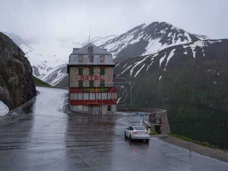 Téléchargez les photos : FURKA PASS, SWITZERLAND, JULY 2021: . Spectacular shot of snowy mountains above a hotel in Furkapass on a misty summer day. Modern white car is parked outside a vintage Belvedere hotel in Furkapass. - en image libre de droit