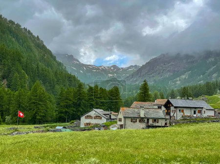 Foto de Scenic view of the rocky Swiss mountains surround the quiet village of Simplon on a cloudy summer day. Picturesque mountain ranges tower above a lush green valley in Switzerland. Cloudy day in Simplon - Imagen libre de derechos