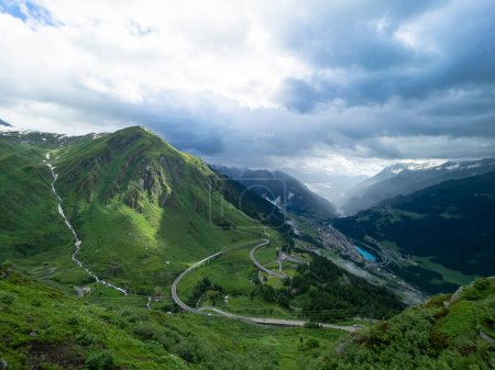 Téléchargez les photos : AERIAL: San Gottardo mountain pass road leads down the lush green valley and toward a remote town in the Swiss Alps. Spectacular view of a massive green gorge in the scenic mountains of Switzerland. - en image libre de droit