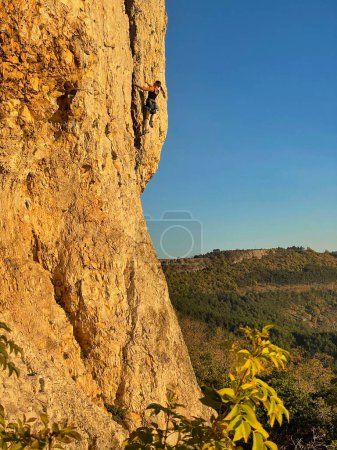 Foto de Athletic young woman climbs a rocky wall in Crni Kal on a sunny autumn morning. Fearless Caucasian female tourist goes top rope climbing up a famous wall in rural Slovenia at fall sunrise. - Imagen libre de derechos