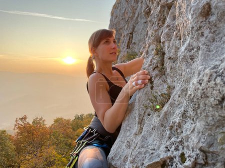 Téléchargez les photos : Young Caucasian woman with taped fingers is rock climbing at golden fall sunrise. Active female tourist climbs up a challenging cliff in the sunlit Slovenian Karst countryside. - en image libre de droit