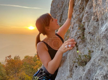 Foto de Active female tourist climbs up a challenging cliff in the sunlit Slovenian Karst countryside. Young Caucasian woman with taped fingers is rock climbing at golden fall sunrise. - Imagen libre de derechos