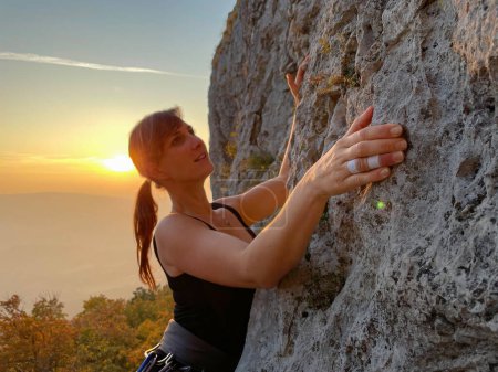 Foto de Happy female climber scales a challenging cliff in sunlit Crni Kal, Slovenia on a sunny autumn evening. Athletic woman climbs up a rocky wall in the idyllic fall countryside. - Imagen libre de derechos