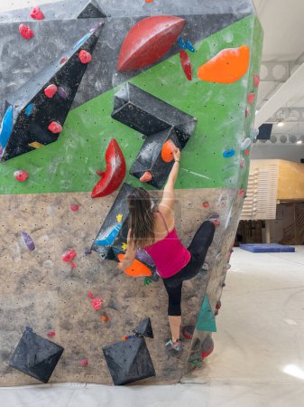 Téléchargez les photos : Athletic female climber navigates the colorful holds while scaling a challenging boulder route at a training hall. Nimble young woman does the splits while climbing indoors at a bouldering center. - en image libre de droit