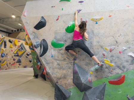 Téléchargez les photos : Nimble young woman does the splits while climbing indoors at a bouldering center. Athletic female climber navigates the colorful holds while scaling a challenging boulder route at a training hall. - en image libre de droit