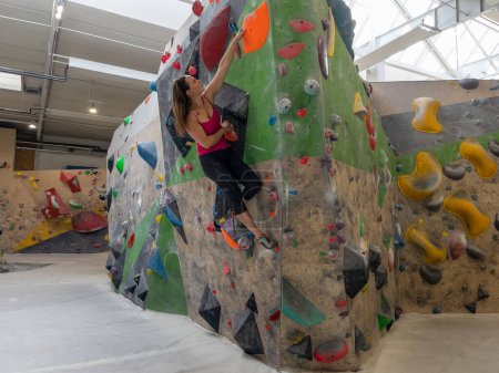 Téléchargez les photos : Nimble young woman does the splits while climbing indoors at a bouldering center. Female climber navigates the colorful holds while scaling a challenging boulder route at a training hall. - en image libre de droit