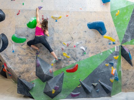 Photo for Agile climber navigates the colorful holds while scaling a challenging boulder route at a training hall. Nimble young woman does the splits while climbing indoors at a bouldering center. - Royalty Free Image