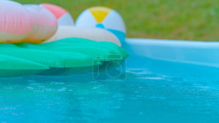 Foto de CLOSE UP, DOF: Crystal clear rain droplets fall into the empty turquoise water and colorful floaties. Cinematic shot of a backyard pool filled with floaties getting caught in an autumn rainstorm. - Imagen libre de derechos