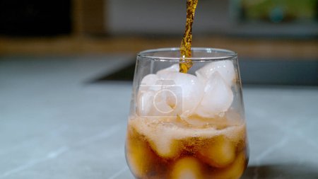 Téléchargez les photos : CLOSE UP, DOF: Bubbling black colored soda gets poured over the ice cubes melting in the elegant glass sitting on the marble kitchen countertop. Dark fizzy drink trickles into a glass full of ice. - en image libre de droit