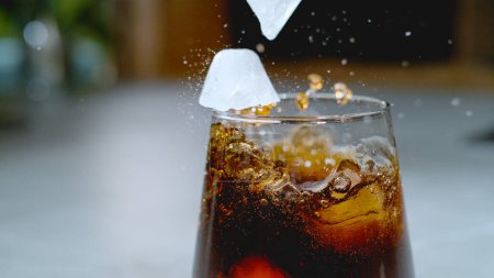 Foto de CLOSE UP, DOF: Odd shaped ice cubes fall into a glass full of fizzy coke. Detailed shot of black colored soda splashing after pieces of ice fall into a glass. Ice falling into bubbling soft drink. - Imagen libre de derechos