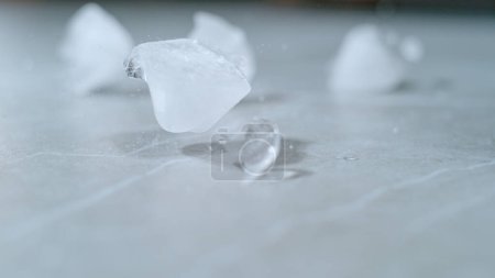 Téléchargez les photos : MACRO, DOF: Melting pieces of ice get scattered across the empty dining table. Odd shaped ice cubes fall and bounce around the marble countertop. White ice cubes are bouncing around the counter. - en image libre de droit