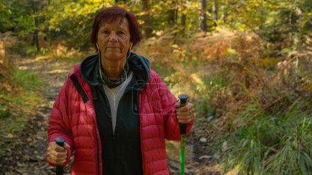 Foto de Smiling female retiree unwinds by listening to a podcast and exploring the colorful autumn forest. Elderly Caucasian woman listens to music while trekking down a forest trail. - Imagen libre de derechos