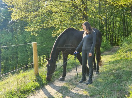 Téléchargez les photos : Young Caucasian woman watches her chestnut horse graze on grass on the side of the forest trail while exploring the beautiful countryside on a sunny autumn day. Horseback rider walking with her mare. - en image libre de droit