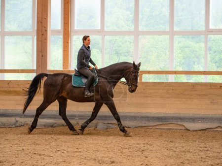 Foto de Young Caucasian woman is horseback riding indoors as training for a competition. Female horseback rider trains with her big chestnut horse at an indoor riding hall. Happy girl is riding a stallion - Imagen libre de derechos