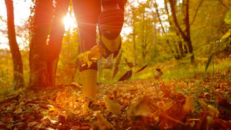 Foto de LENS FLARE, CLOSE UP, LOW ANGLE: Unrecognizable happy woman wearing yellow boots runs in the fall forest turning leaves. Energetic girl in rubber boots kicks up fallen leaves while jogging in woods - Imagen libre de derechos