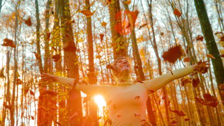 Téléchargez les photos : CLOSE UP, LENS FLARE: Carefree girl exploring the woods plays with dry leaves. Happy Caucasian woman trekking in the autumn colored woods throws dry fallen leaves high in the air at golden sunrise. - en image libre de droit