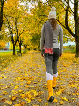 Photo for Woman strolls along a scenic autumn colored avenue in her yellow rubber boots on a cold November afternoon. Young female walks along a walkway crossing the park - Royalty Free Image